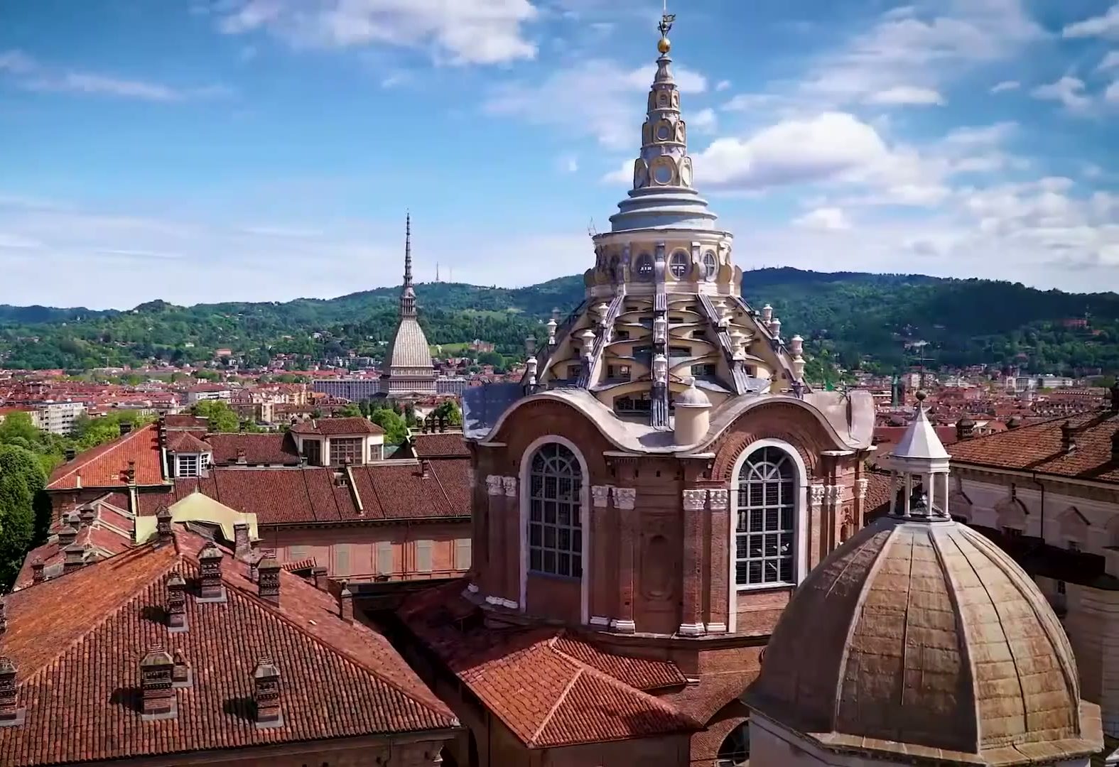 Vista aérea de las cúpulas barrocas y los tejados de Turín, con la Mole Antonelliana y colinas verdes al fondo