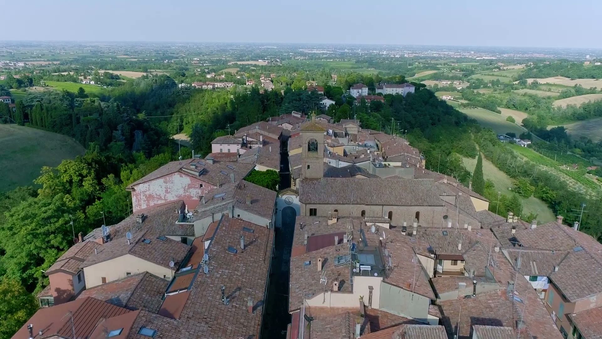 Vista aérea de un pueblo de Emilia-Romaña en lo alto de una colina, con un campanario entre ondulados campos verdes