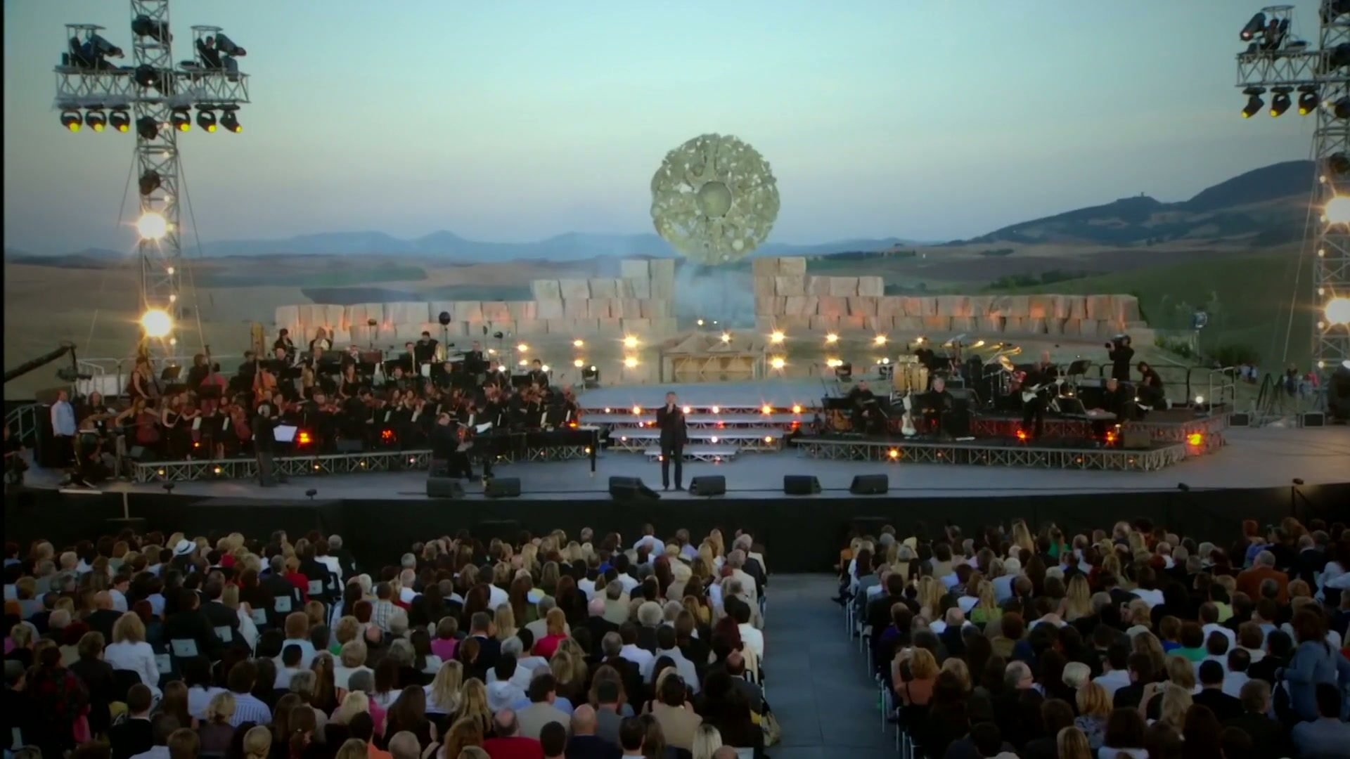 An open-air orchestral concert stage and audience surrounded by rolling Tuscan hills at dusk