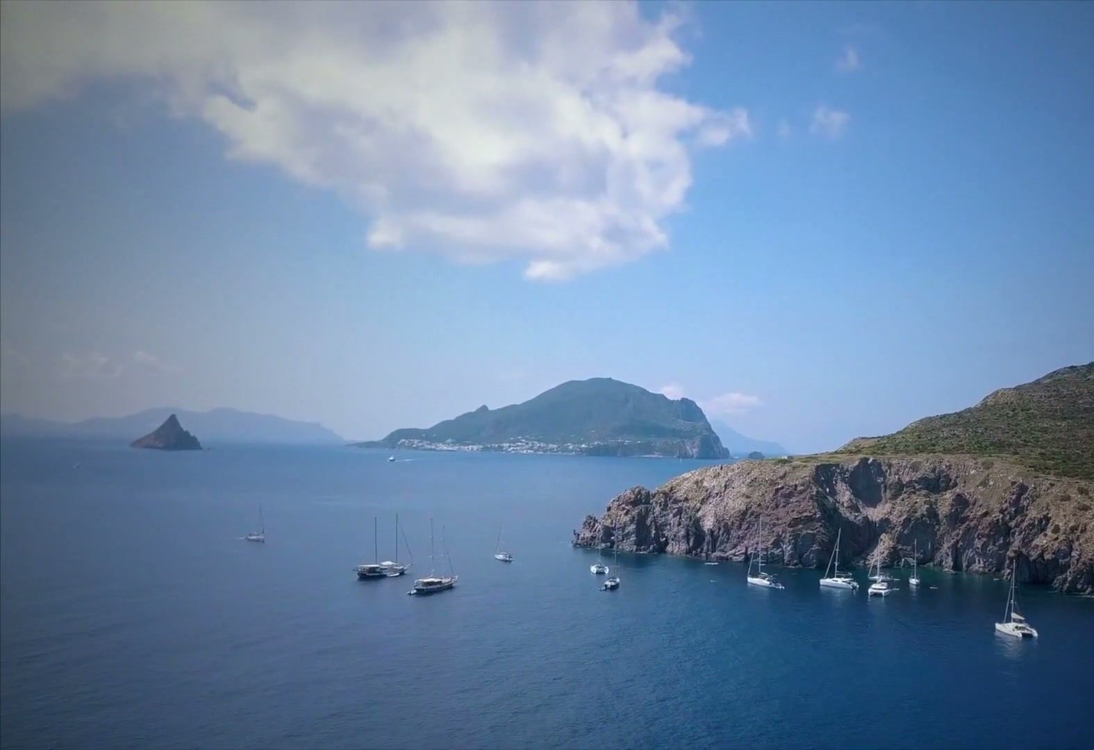 Sailboats anchored beside a rocky Aeolian coastline with volcanic islands beyond